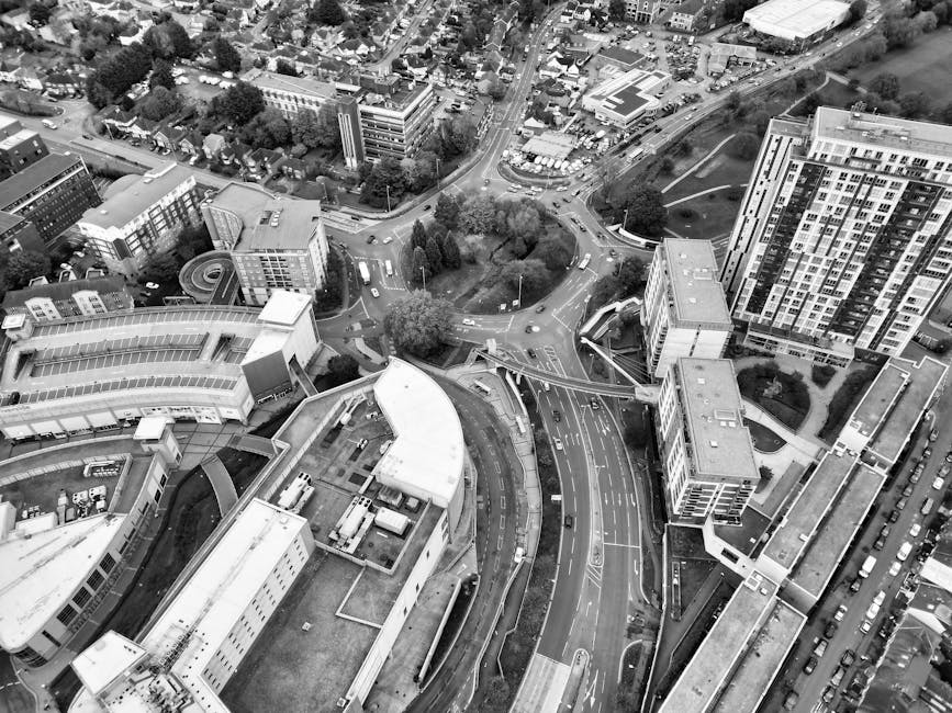 An aerial black-and-white photograph capturing a busy urban area with a mix of modern office buildings, high-rise residential towers, and smaller structures. Prominent in the foreground is a curved commercial or public building with a flat roof, surrounded by multiple vehicles parked along adjacent streets and loading bays. The roads intersect with a roundabout and are lined with trees, indicating a landscaped town centre environment. In the background, there are green open spaces, possibly parks or sports fields, alongside additional buildings and parking lots. The scene reflects dense city infrastructure typical of a town centre, with clear visibility of vehicular and pedestrian movement. The image relates to rubbish removal services by highlighting the scale of urban waste management challenges faced in such busy areas, consistent with independent rubbish clearance solutions provided by Rubbish Clearance Surrey, serving the Surrey region.