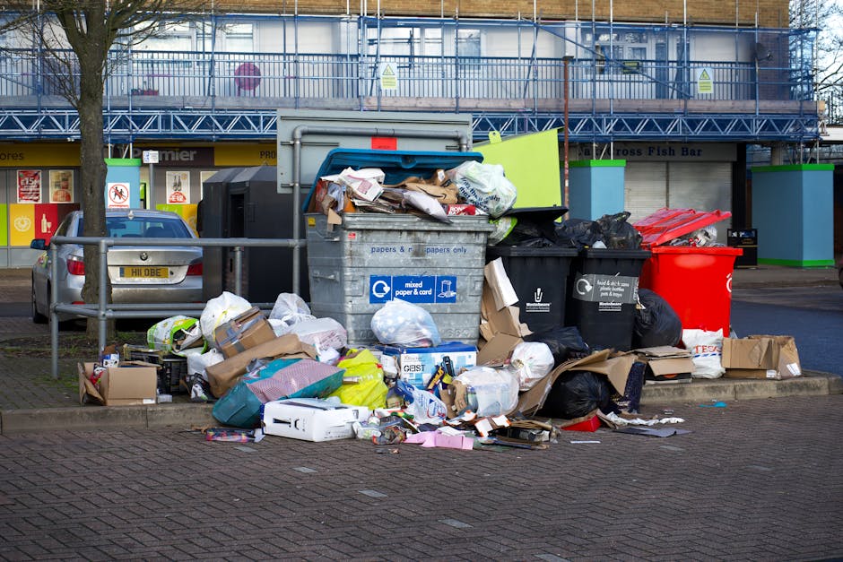 A large outdoor collection of mixed household rubbish and waste materials piled around multiple bins and bags on a paved area beside a parking space. The waste includes cardboard boxes, plastic bags, paper, and assorted packaging, some of which are partially torn or overflowing. The bins are of various types and colors, with a grey bin designated for mixed paper and card, a black recycling bin, and a red bin possibly for general waste. The waste is scattered on the ground in front of and around the bins, with some items leaning against the bins and others spilling onto the pavement. In the background, there is a commercial building with signage, a metal security fence, a silver car parked nearby, and trees along the scene. The surroundings suggest a designated waste collection point that may be managed by a private rubbish removal service, such as Rubbish Clearance Surrey, for a commercial or residential area in Surrey. The scene is lit by natural daylight with a slightly overcast sky, highlighting the cluttered and unmanaged appearance of the waste storage area.