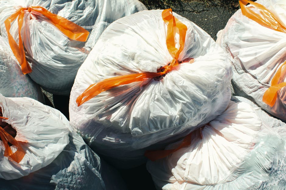 A group of large white plastic rubbish bags tightly sealed with orange ties, filled with mixed waste material, are piled together on an outdoor surface. The bags appear to be made of durable, semi-transparent plastic, allowing some of the contents to be faintly seen through the material. They are arranged in a clustered manner, with some bags leaning against each other and others lying flat, suggesting recent collection or temporary storage for a private rubbish removal service. The background shows a dark, paved or asphalt area, typical of a driveway or waste collection point, with no other objects or environmental features visible. The scene is illuminated with natural daylight, casting soft shadows and highlighting the crinkled texture of the plastic bags, emphasizing their bulk and the secure sealing method used. This visual depicts a typical scenario of domestic or commercial waste awaiting pickup by a professional waste management company like Rubbish Clearance Surrey, which offers alternative waste disposal solutions outside local authority provisions for rubbish removal.