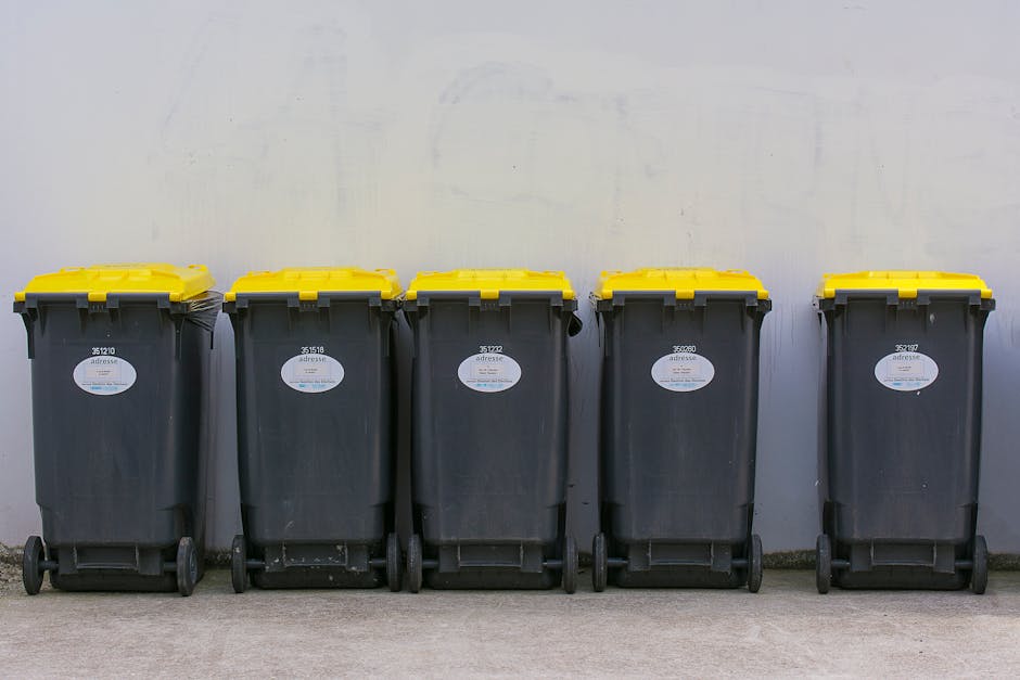 A large outdoor collection of mixed household rubbish and waste materials piled around multiple bins and bags on a paved area beside a parking space. The waste includes cardboard boxes, plastic bags, paper, and assorted packaging, some of which are partially torn or overflowing. The bins are of various types and colors, with a grey bin designated for mixed paper and card, a black recycling bin, and a red bin possibly for general waste. The waste is scattered on the ground in front of and around the bins, with some items leaning against the bins and others spilling onto the pavement. In the background, there is a commercial building with signage, a metal security fence, a silver car parked nearby, and trees along the scene. The surroundings suggest a designated waste collection point that may be managed by a private rubbish removal service, such as Rubbish Clearance Surrey, for a commercial or residential area in Surrey. The scene is lit by natural daylight with a slightly overcast sky, highlighting the cluttered and unmanaged appearance of the waste storage area.