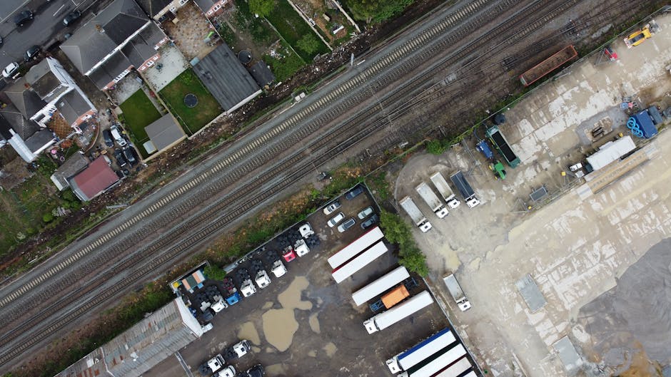 An aerial view showing a variety of parked vehicles, including cars and trucks, arranged on a gravel and paved lot adjacent to a railway line. The site contains numerous white and blue storage containers, trailers, and scattered equipment, some of which appear to be used for waste or materials storage. Puddles of water and muddy patches are visible on the uneven ground, suggesting recent rainfall. To the left, there is a residential area with houses and small gardens, separated from the industrial yard by a railway track running horizontally across the image. The background features greenery and trees, with the overall scene representing an industrial or commercial space involved in logistics, storage, or waste handling activities, consistent with independent rubbish clearance services. The environment appears well-lit under natural daylight, with no visible signage or branding in the image.